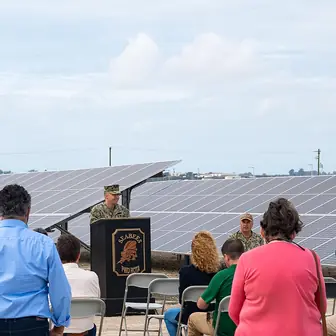 A speaker at a lectern in front of long rows of photovoltaic panels